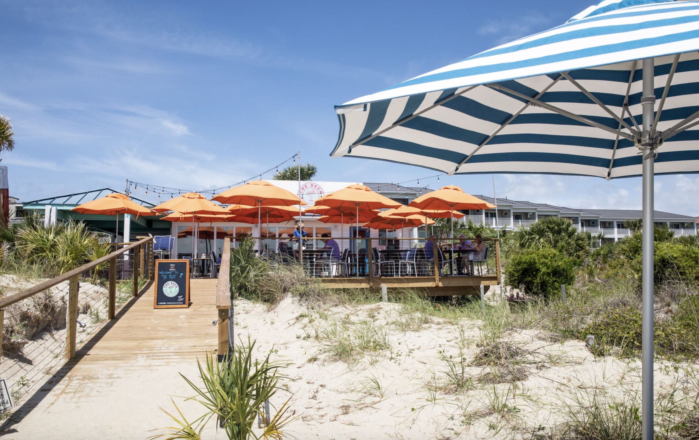A beachside restaurant with outdoor seating under striped umbrellas, a wooden deck leading to the entrance, and a sign welcoming guests.