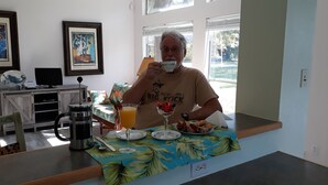 Un homme est assis à une table recouverte d’une nappe aux motifs tropicaux, avec une machine à café et un verre de jus d’orange.
