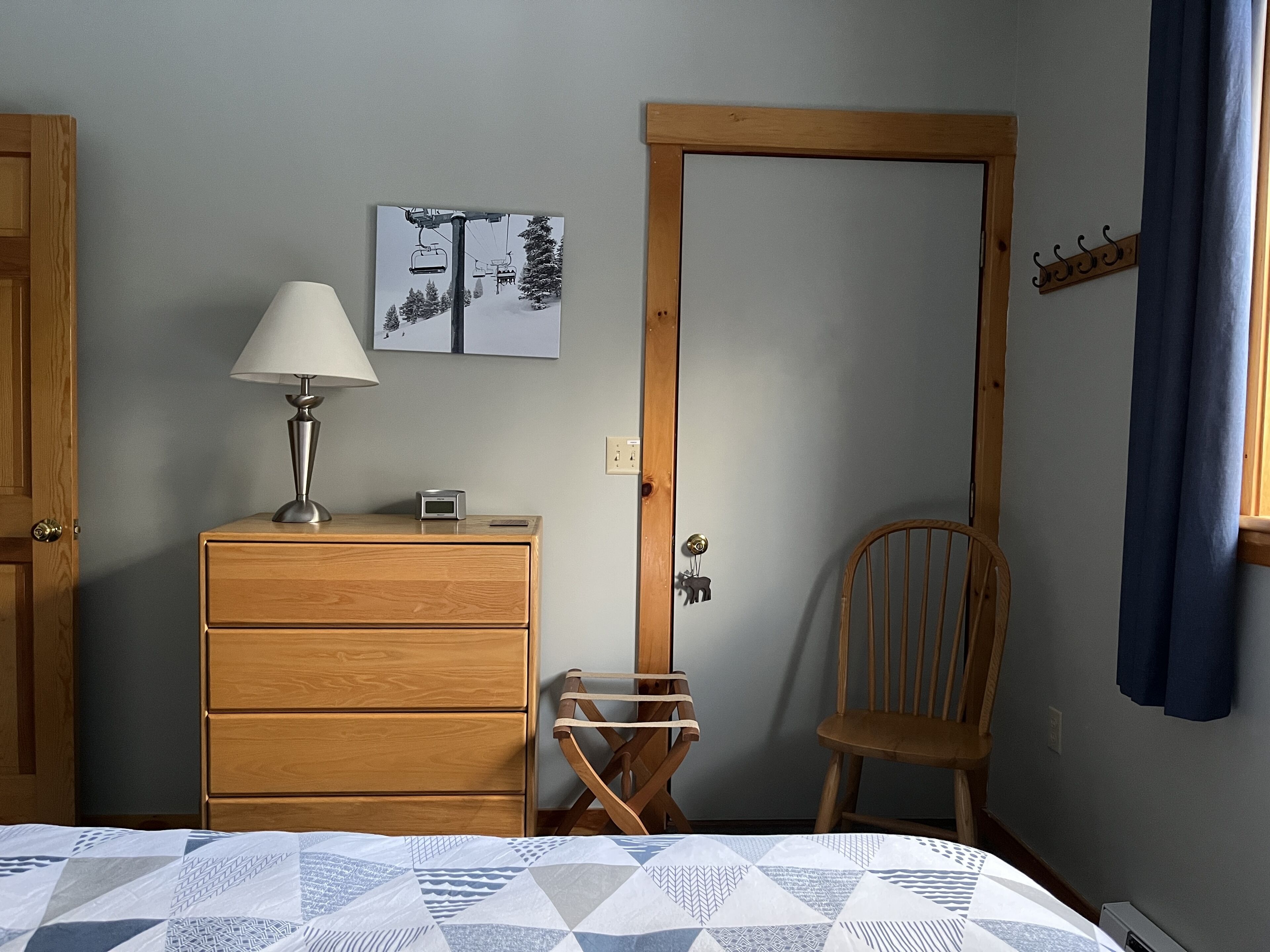 Une chambre avec une commode en bois, une chaise, un lit recouvert d’une courtepointe à motifs, une lampe fixée au mur et une image représentant un télésiège.
