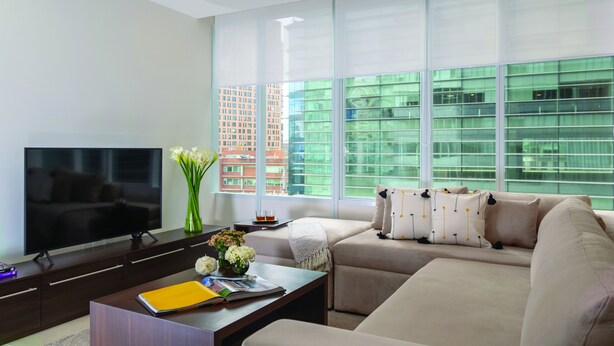 A modern living room with a large flat-screen TV, a sofa, a coffee table, and a window with a view of buildings.