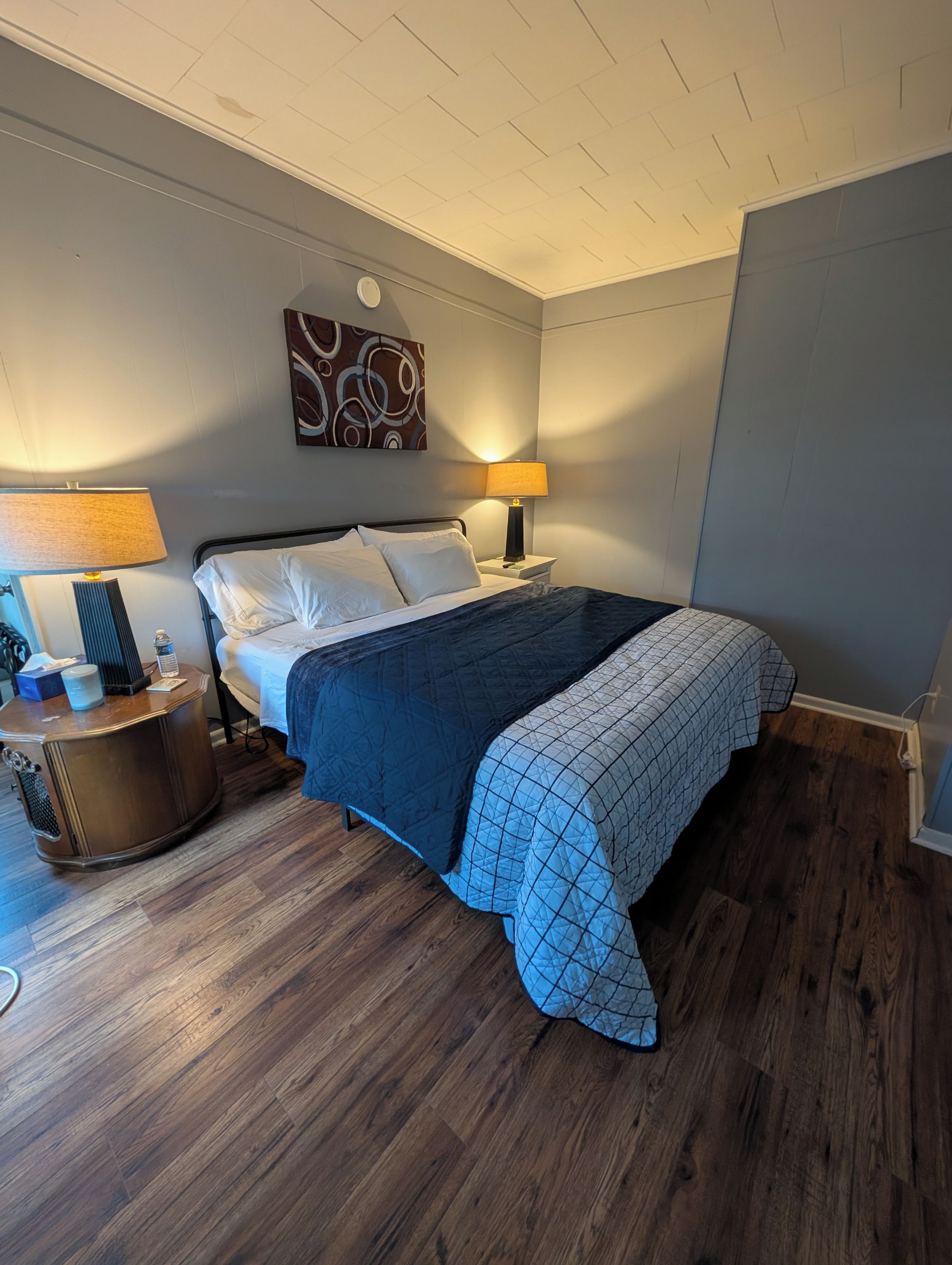 A hotel room with a bed, two bedside tables, a lamp, a wall clock, and a painting.
