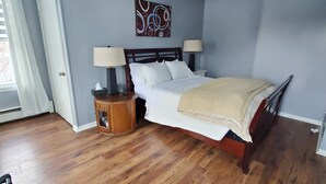 Un dormitorio con piso de madera, una cama con ropa de cama blanca, dos lámparas de noche, una mesita de noche y una obra de arte en la pared.