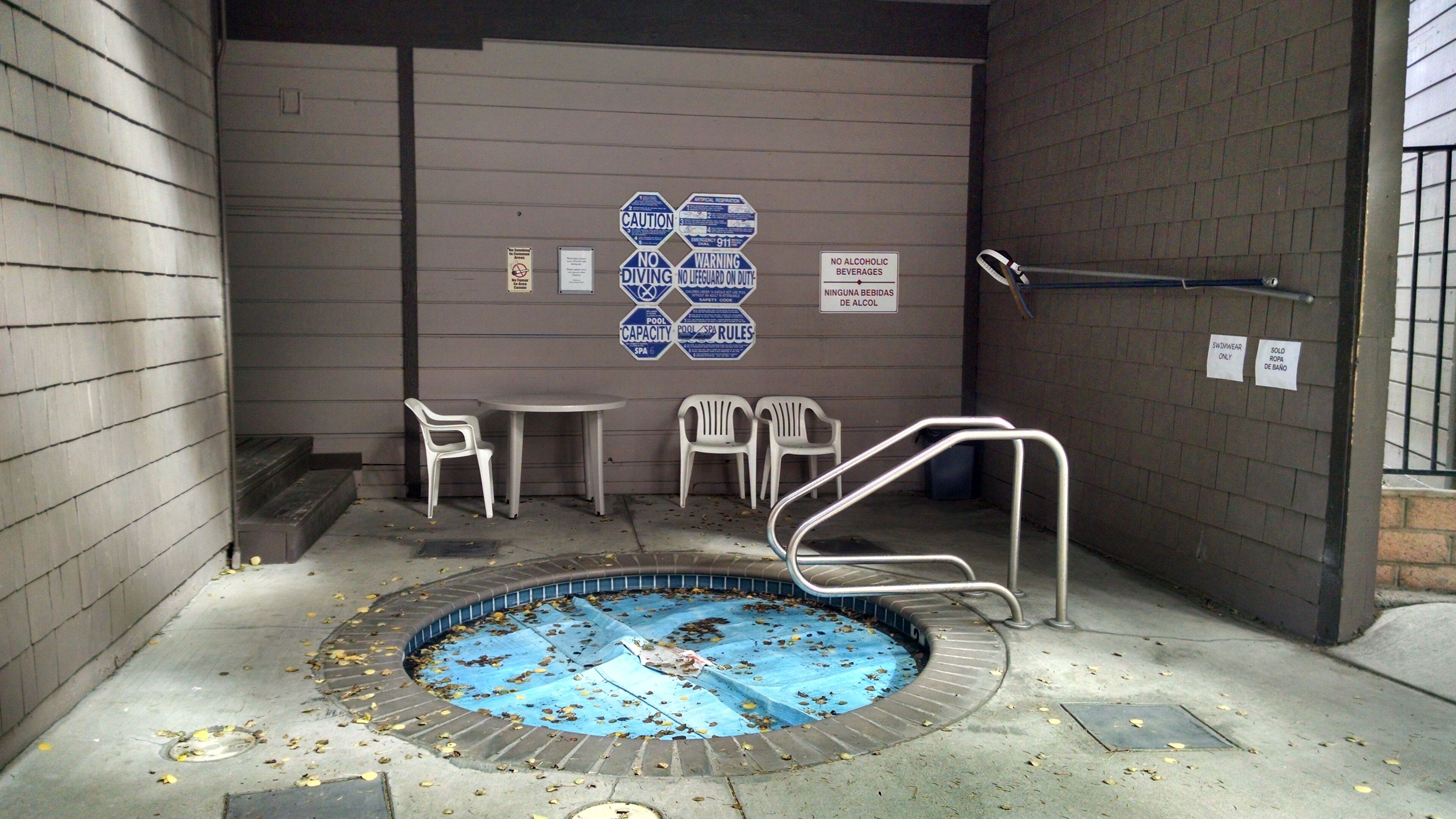 A small indoor pool with a blue pool cover, white plastic chairs, and a metal pool ladder.