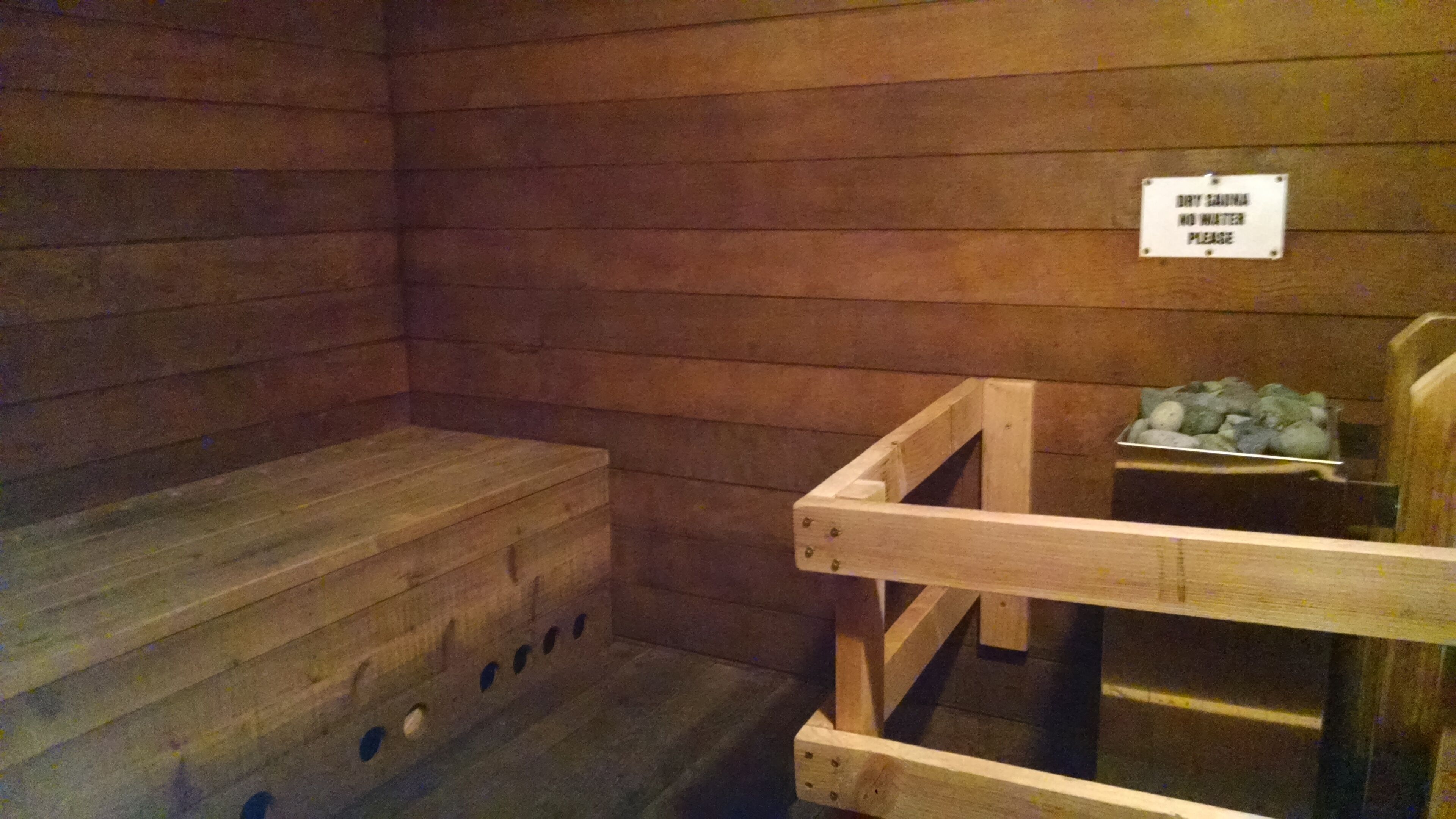 A sauna room with wooden walls and benches. A sign on the wall reads "DRY SAUNA NO WATER PLEASE".