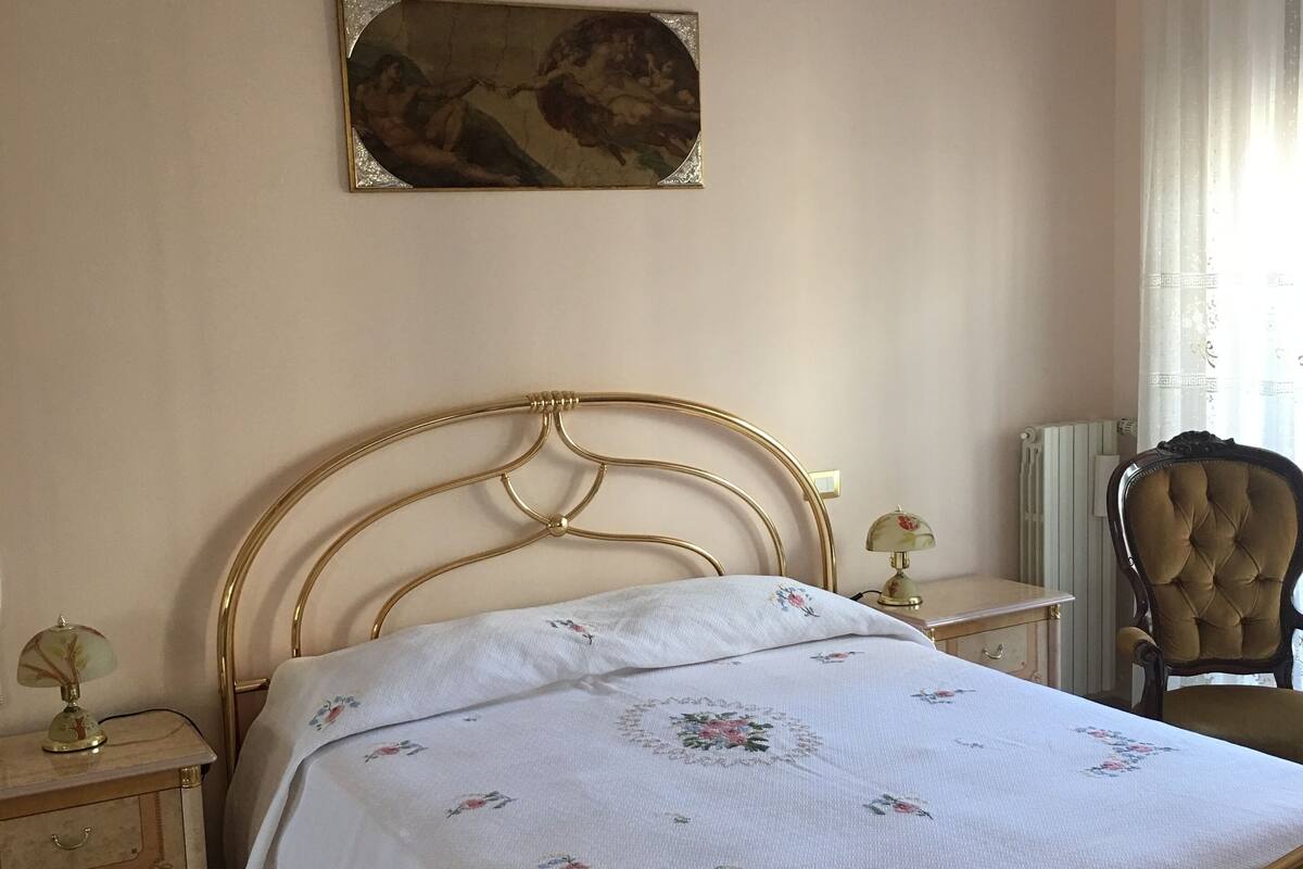 Un dormitorio con cama, mesita de noche, silla, lámpara y un cuadro en la pared.