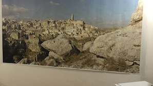 Quarto com uma grande pintura de paisagem urbana na parede, uma pequena mesa com etiqueta de nome e um armário branco.
