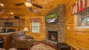 A cozy cabin interior with a stone fireplace, wooden walls, a television mounted above it, a kitchen area with a microwave, and a patterned sofa. - Heavenly View – Romantic Pigeon Forge Cabin with Hot Tub, Jacuzzi & Pool Table (Sevierville)
