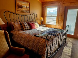 Um quarto aconchegante com estrutura de cama de metal, roupa de cama estampada, uma cadeira, mesa de cabeceira com abajur e uma janela com persianas.