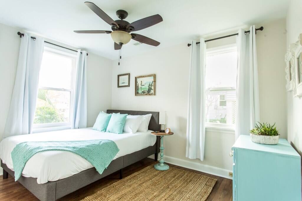 A bedroom with a bed, a ceiling fan, a bedside table, a small plant, and a window with curtains.