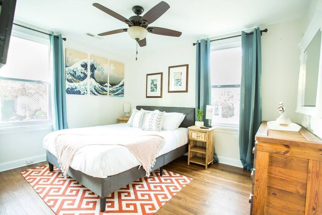 A bedroom with a bed, a ceiling fan, a wooden nightstand, a dresser, and artwork on the wall.