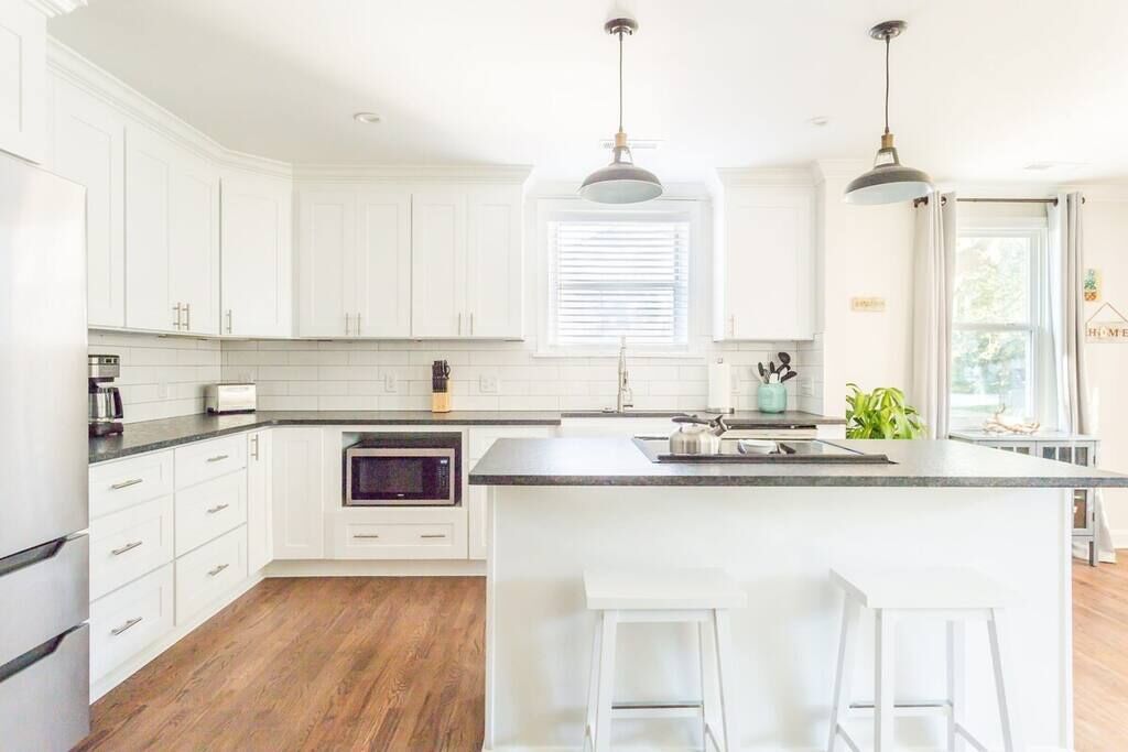 Una cocina moderna con armarios blancos, una isla central con taburetes, horno empotrado e iluminación colgante.