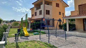 Um parquinho com balanços e escorrega, cercado por uma área residencial com um edifício de dois andares.