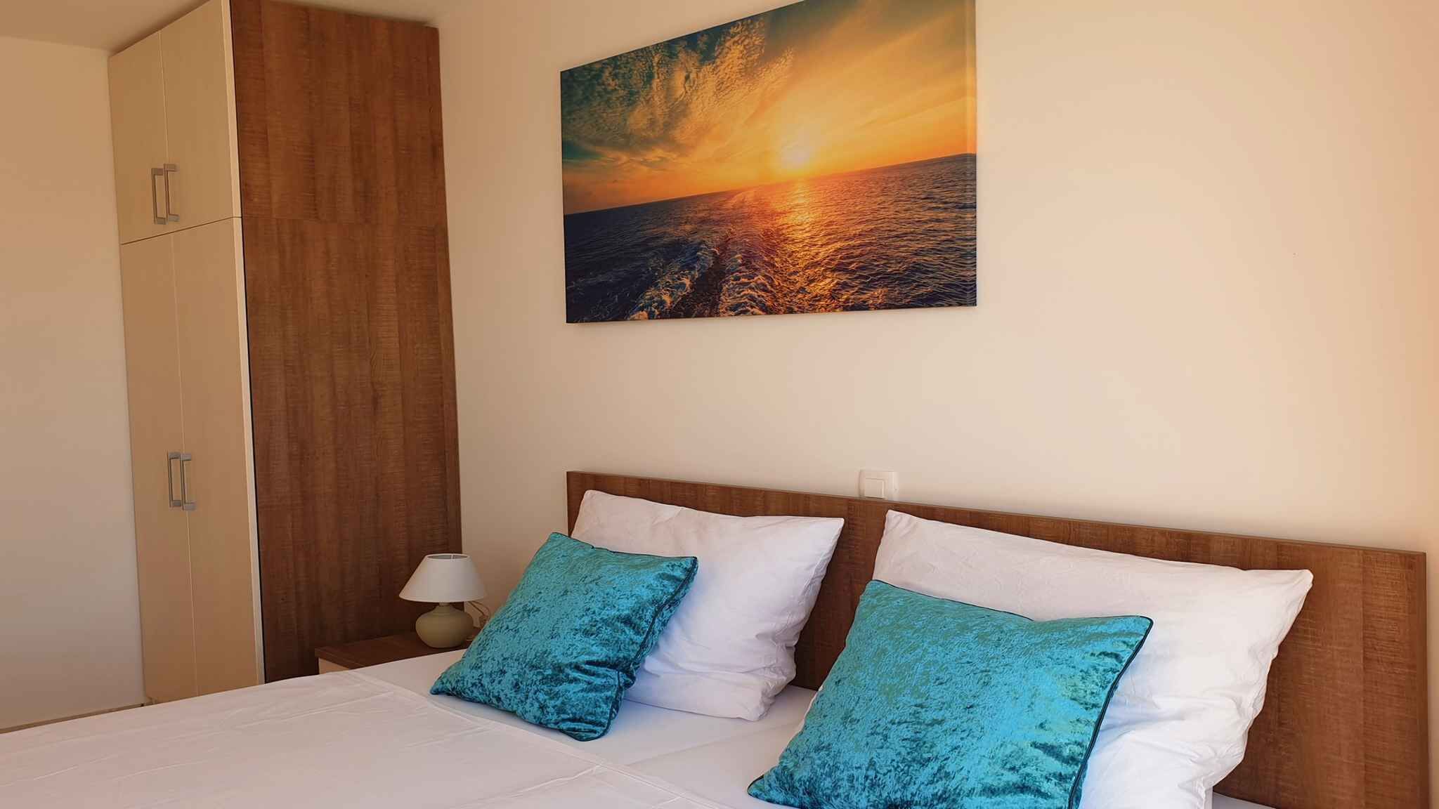 Ein Hotelzimmer mit einem Bett, zwei Kissen, einem Nachttisch, einer Lampe und einem Gemälde, das einen Sonnenuntergang über dem Meer zeigt.