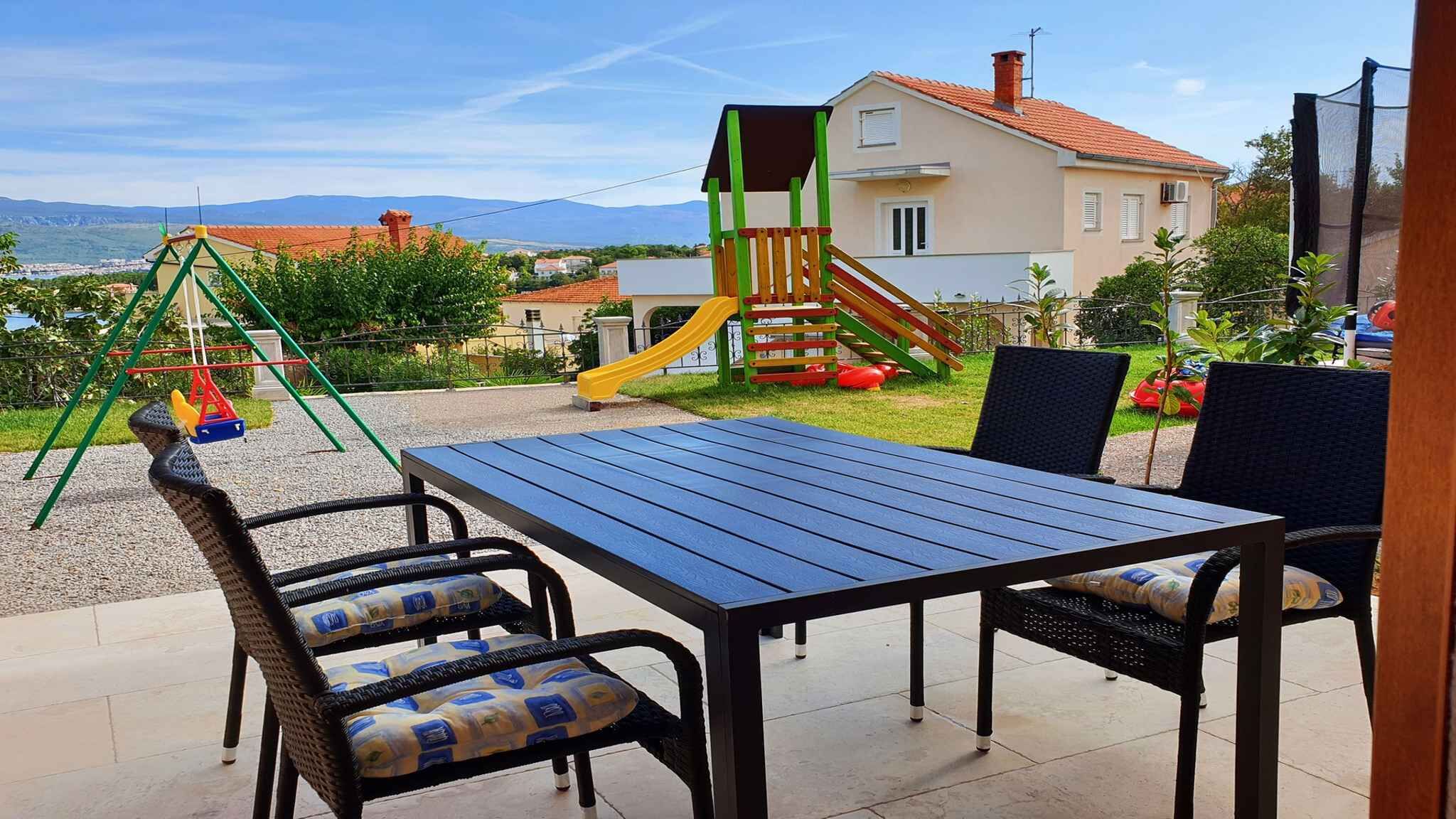 Eine Terrasse im Freien mit Tisch und Stühlen, ein Spielbereich für Kinder und ein Blick auf ein Haus sowie Berge im Hintergrund.