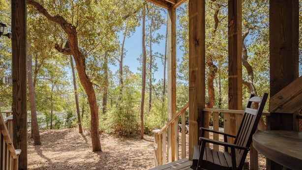 Una terraza de madera con una silla y una mesa, rodeada de árboles y un cielo despejado.