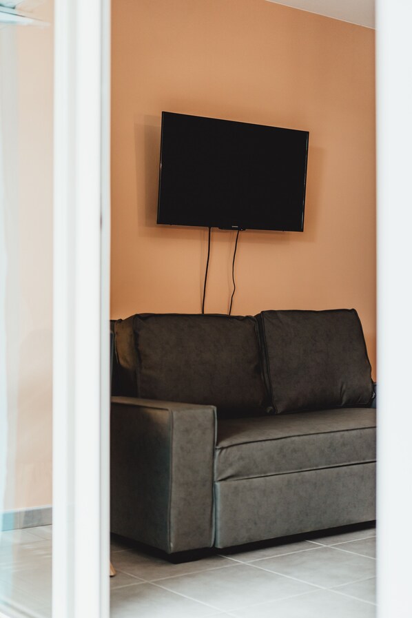 A modern living room with a flat-screen TV mounted on the wall, a grey sofa with cushions, and a tiled floor. - Magnificent F1 Apartment, The Coral Garden, in the Heart of Saline les Bains (La Saline-Les-Bains)