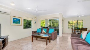 A spacious living area with a sofa, coffee table, and a TV stand. There are large windows and a balcony with a view of greenery.