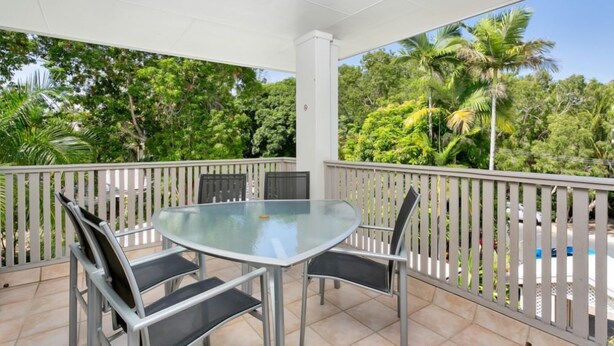 A covered outdoor area with a glass table and chairs, surrounded by a white railing, overlooking a lush green landscape.