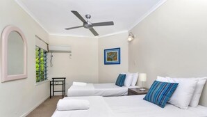 A hotel room with two beds, a ceiling fan, a window with a view of greenery, a framed picture on the wall, and a small table with a lamp.