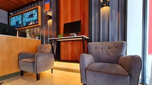Un moderno vestíbulo de hotel con un mostrador de recepción de madera, dos sillones grises, un televisor de pantalla plana y un gran ventanal con cortinas.