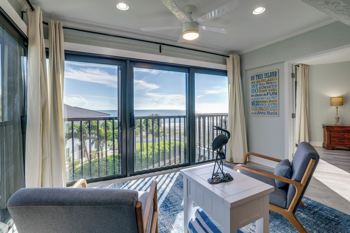 A room with a large sliding glass door leading to a balcony, a ceiling fan, a white desk, two chairs, and a wall with a poster.