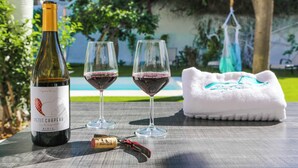 Une bouteille de vin Petit Chapeau, deux verres de vin rouge, un tire-bouchon et une serviette blanche sur une table à l’extérieur.