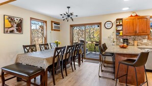A dining area with a wooden table and chairs, a kitchen with wooden cabinets, and a balcony with a view of trees. - Cozy Creekfront House with Sauna (Bushkill)