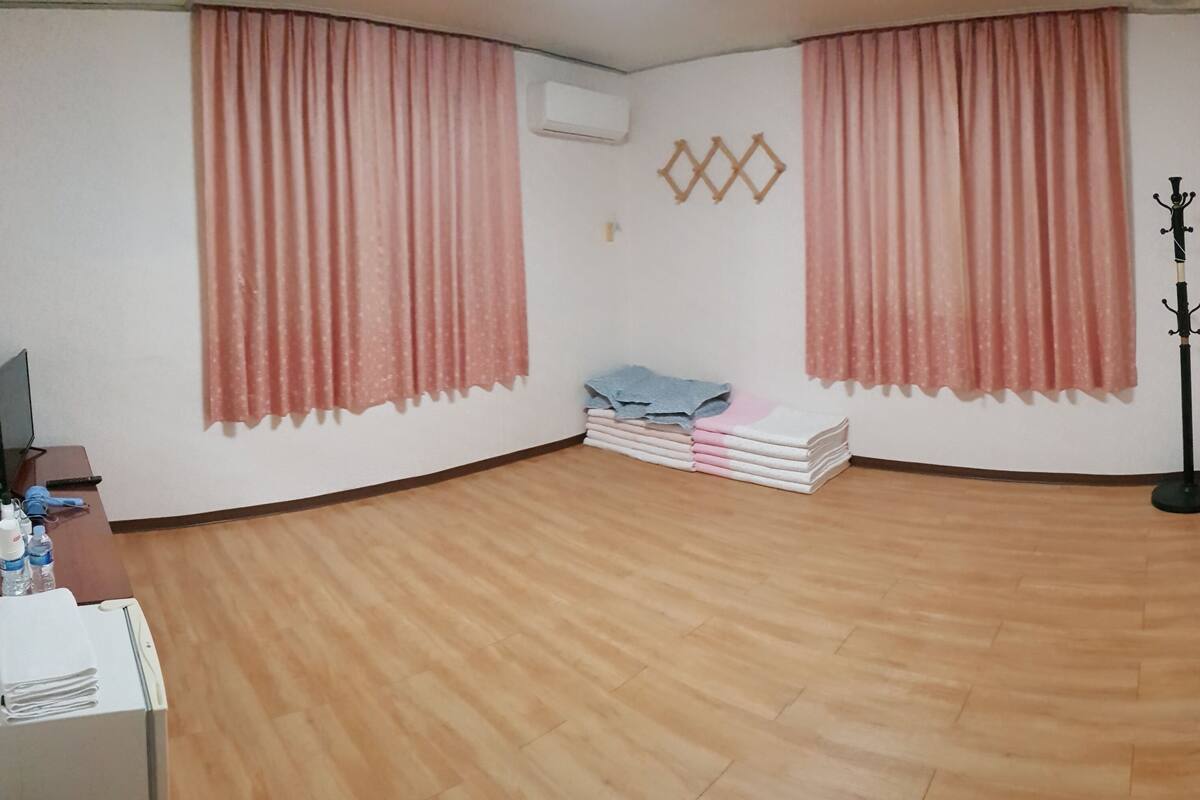 Ein Hotelzimmer mit Holzboden, rosafarbenen Vorhängen, einem Kleiderständer und einem Stapel gefalteter Handtücher.