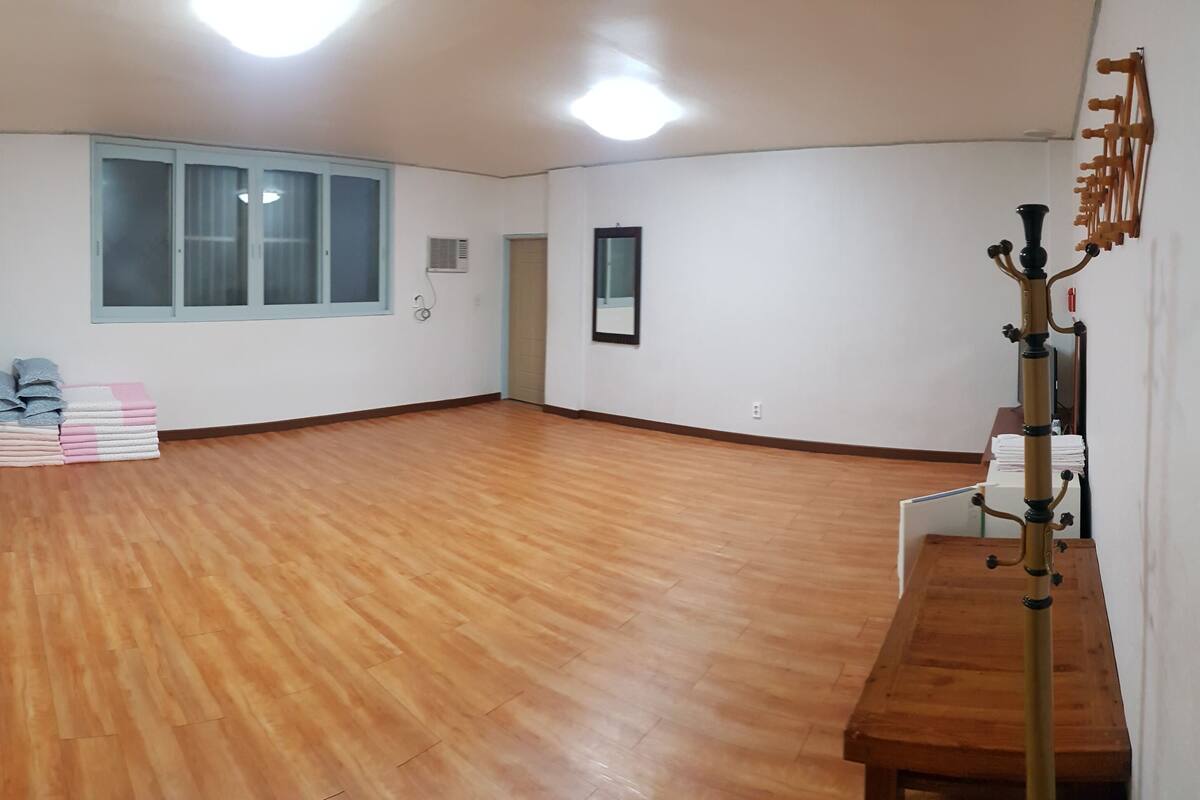 Ein geräumiges Zimmer mit Holzboden, einem großen Fenster mit Jalousien, einem Holztisch und einem an der Wand befestigten Regal.