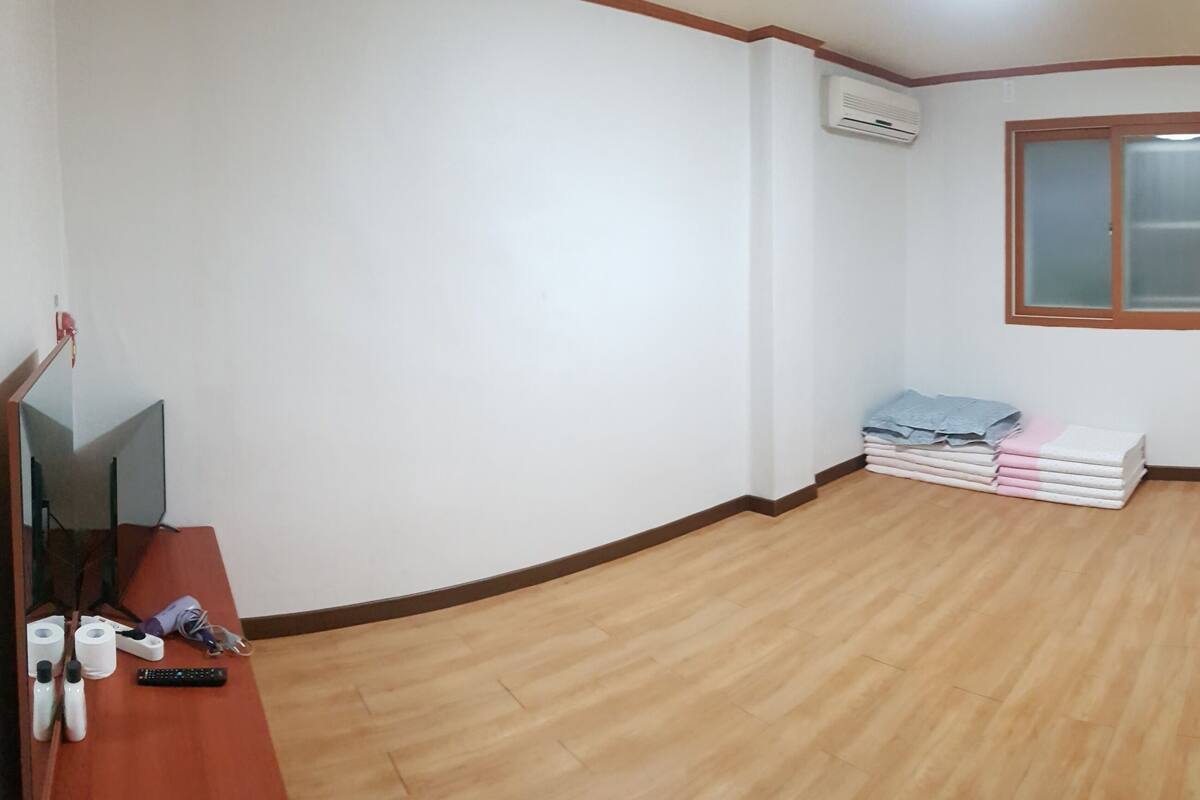 Ein Zimmer mit Holzboden, einem Fernseher auf einem Schrank, einem Fenster und einer Wandklimaanlage.