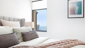A modern bedroom with a large window, a bed with multiple pillows, and a view of the sea.