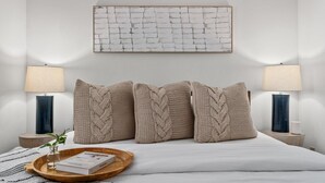 Ein Schlafzimmer mit einem Bett, zwei Nachttischlampen, einem an der Wand befestigten abstrakten Kunstwerk und einem Nachttisch mit einem Buch und einem Glas.