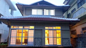 A traditional Japanese house with a tiled roof, two large windows with sliding doors, and a small garden area in front. - Mahoroba (Motobu)