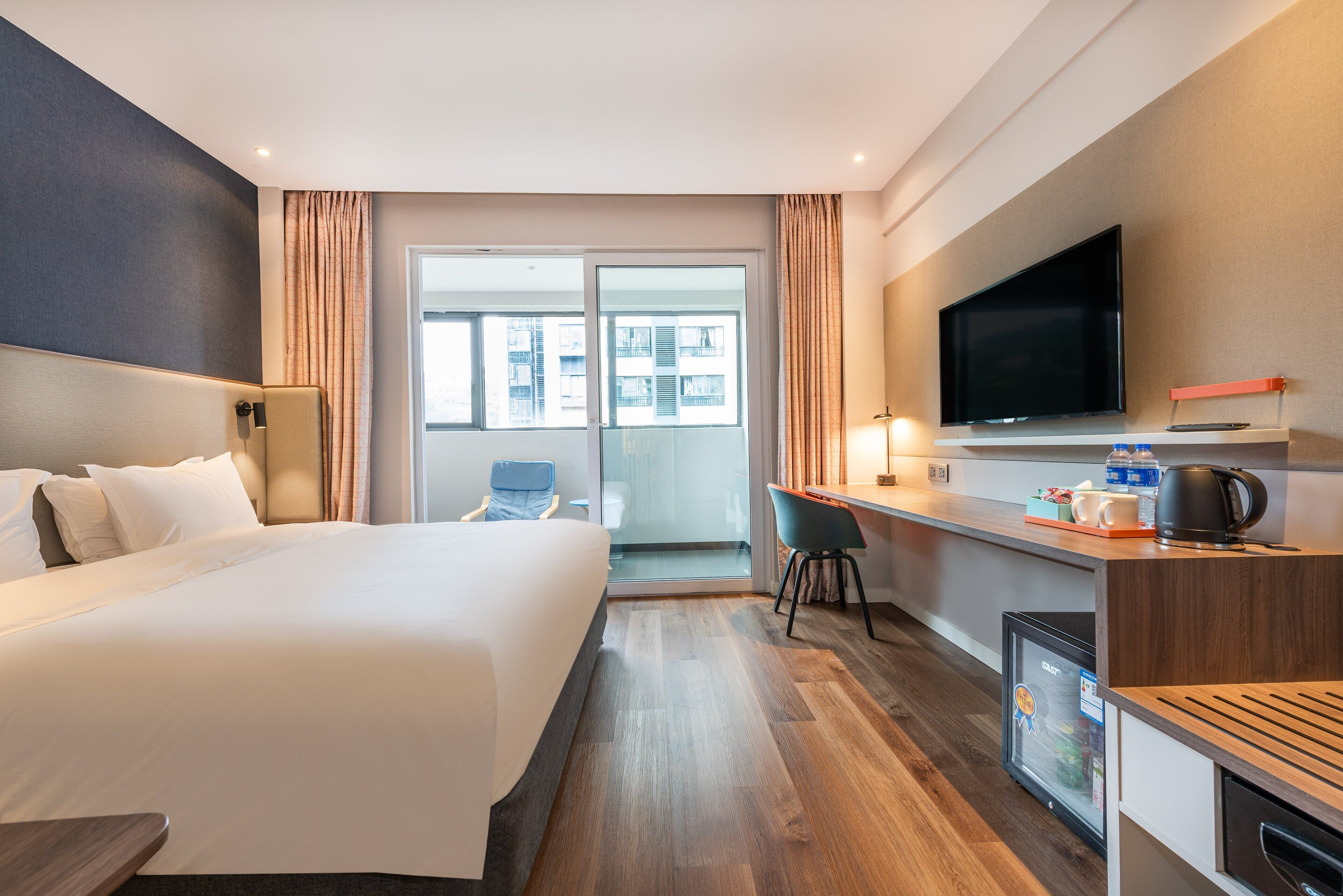 Ein modernes Hotelzimmer mit einem großen Bett, einem Schreibtisch mit Flachbildfernseher, einer Minibar und einer Sitzecke mit einem Sessel.