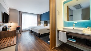 Ett hotellrum med ett skrivbord i trä, en säng, en stor spegel och ett badrum med handfat och spegel.