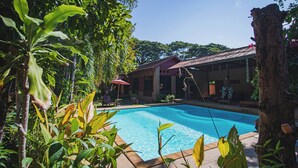 A swimming pool surrounded by lush greenery, with a building in the background. - Joy's House (Chiang Mai)
