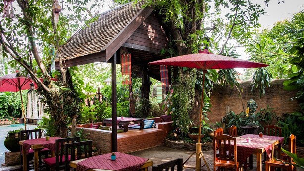 An outdoor patio with wooden tables and chairs, covered by red umbrellas, surrounded by lush greenery and a brick wall. - Joy's House (Chiang Mai)