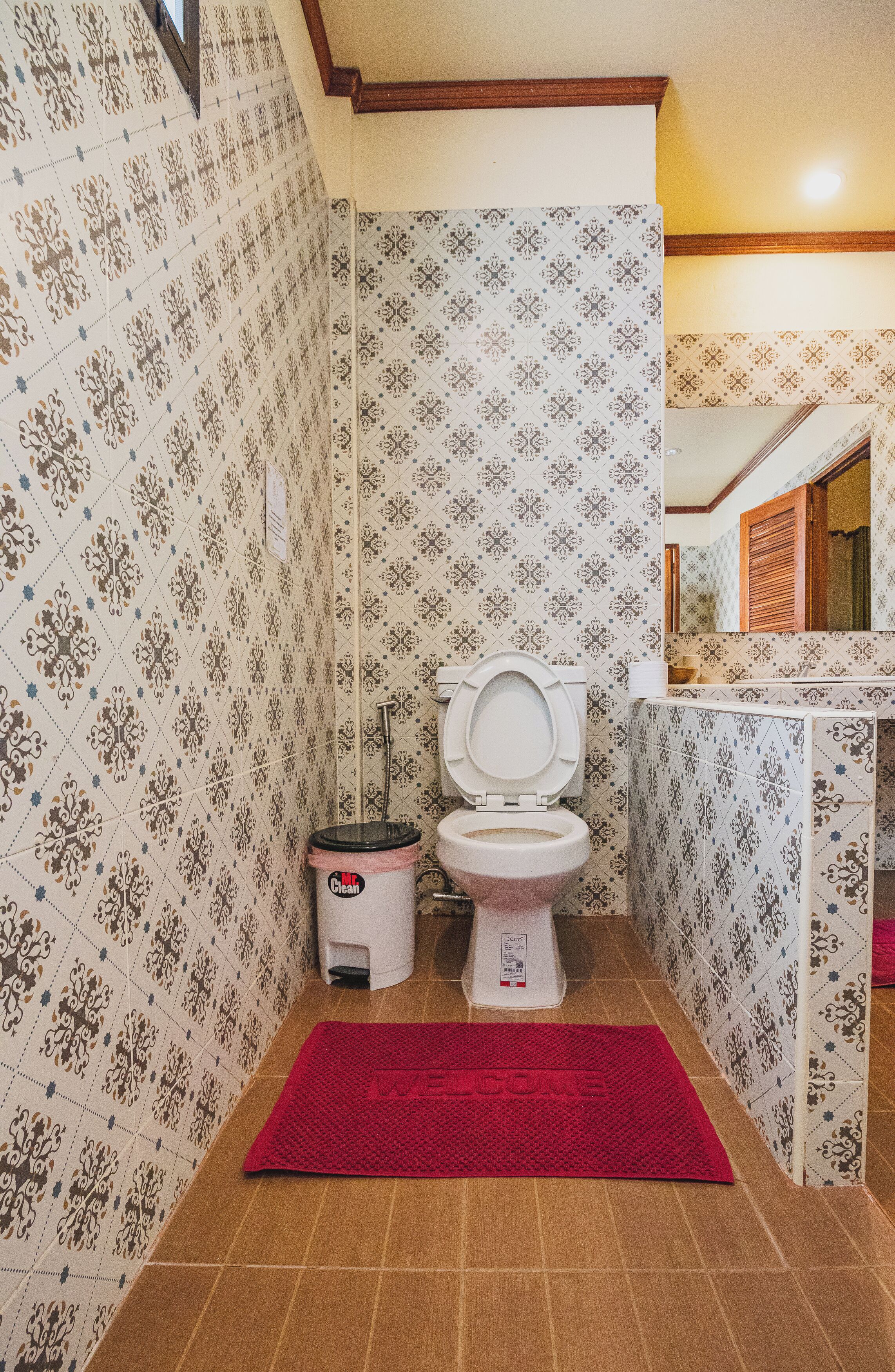 Een badkamer met patroontegels, een toilet, een prullenbak en een rode welkomstmat.