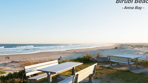Una playa con bancos y mesas de picnic, vista despejada del océano y el texto "Birubi Beach - Anna Bay - TheBay".