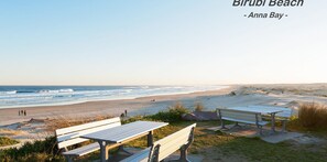 Une plage avec des bancs et des tables de pique-nique, une vue dégagée sur l’océan, et l’inscription : « Birubi Beach – Anna Bay – TheBay ».