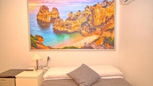 A hotel room with a bed, a desk with a lamp, a suitcase, and a painting of a coastal landscape. - Great apartment with pool in Los Boliches (FUENGIROLA)