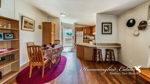 A compact kitchen with wooden cabinets and a central island, a dining area with a table and chairs, and a bookshelf.