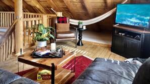 Una habitación estilo loft acogedora, con techo de madera, una hamaca, sofá de cuero, televisor y una mesita con una planta y libros.