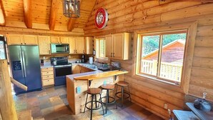 Una cabaña rústica de madera con cocina que incluye refrigerador, estufa y alacenas de madera. Hay un ventanal con vista a una terraza de madera.