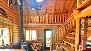 Interior de una cabaña de troncos con una escalera de madera, una zona de cocina y un comedor.