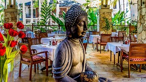 Una estatua de Buda en un restaurante, con mesas y sillas, rodeada de flores rojas y follaje verde.