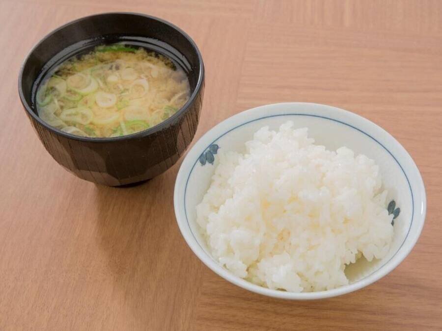 Un tazón de arroz blanco y un tazón de sopa transparente con cebollines sobre una mesa de madera.