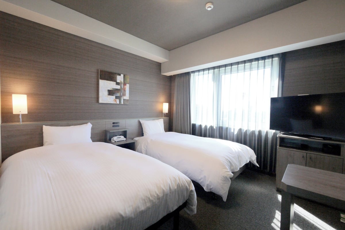 Ein Hotelzimmer mit zwei Betten, einem Flachbildfernseher, einem Schreibtisch und einem Fenster mit Vorhängen.