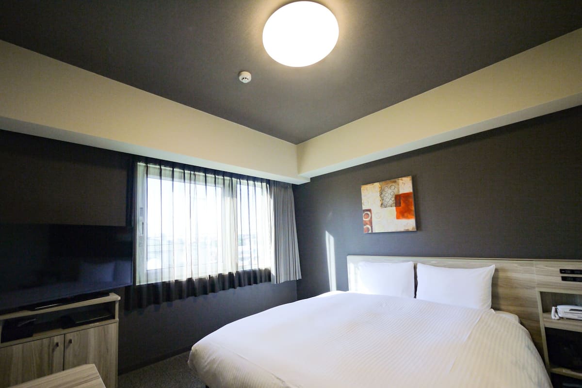 Ein Hotelzimmer mit einem großen Bett, einem Fernseher auf einem Schrank, einem Schreibtisch und einem Fenster mit Vorhängen.