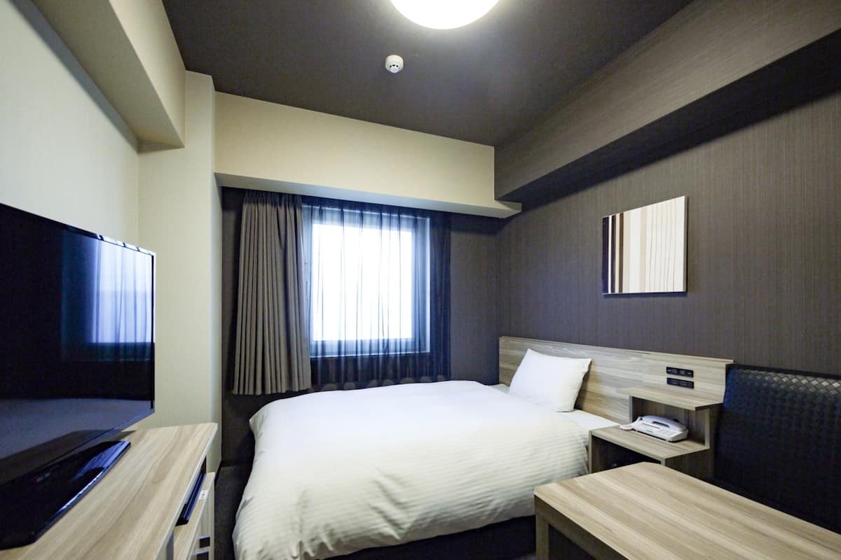 Ein Hotelzimmer mit einem Bett, einem Schreibtisch, einem Fernseher, einem Nachttisch und einem Vorhangfenster.
